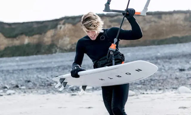 Młody kitesurfer w czarnym piance, doceniając jej właściwości termoizolacyjne, przygotowuje deskę do wodnych szaleństw.