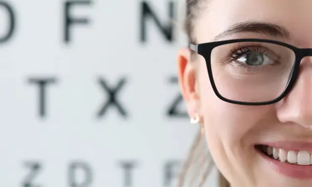 Uśmiechnięta kobieta w okularach na tle tablicy Snellena. Wizyta u optometrysty lub okulisty to ważny krok do dobrego widzenia.