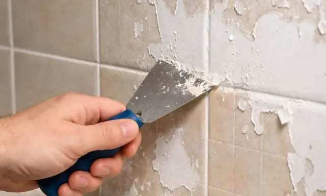 Main tenant enlever peinture sur carrelage avec une spatule. La peinture s'écaille sur les carreaux beiges.