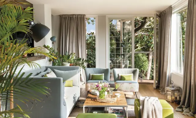 Sofá gris y otomanas verdes crean un ambiente acogedor. Plantas exuberantes y luz natural inundan el salón.