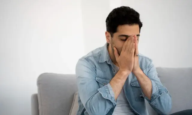 Mann sitzt auf Sofa, Hände vors Gesicht gepresst, Augen geschlossen. Fühlt sich nach dem Rauchen schwindelig.