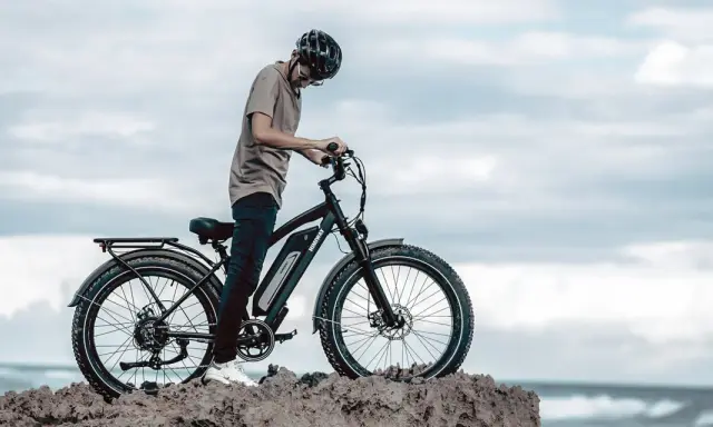 Ein Mann mit Helm steht neben einem Fatbike mit breiten Reifen am Meer. Was ist ein Fatbike? Ein robustes Fahrrad für jedes Gelände.