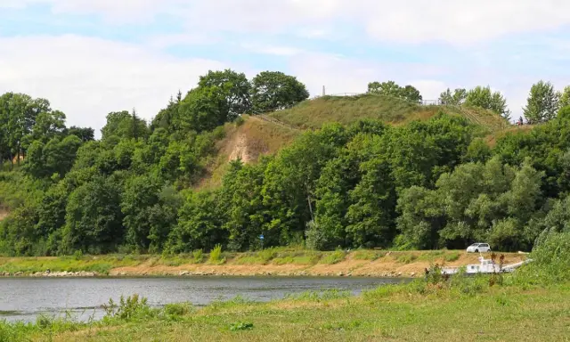 Zielona skarpa nad rzeką, na szczycie której widać ścieżki i ogrodzenie. To góra zamkowa w Drohiczynie, miejsce pełne historii.