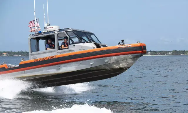 Un bateau du garde côte américain file sur l'eau, créant une gerbe d'écume. Deux membres d'équipage sont visibles à l'intérieur.