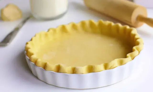 Pâte brisée prête pour la cuisson dans un plat à tarte blanc, avec un rouleau à pâtisserie en arrière-plan.