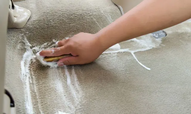 Ręka szoruje szczotką zaplamioną wykładzinę dywanową, tworząc pianę.