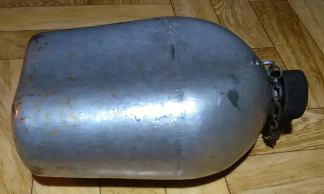 Metalowa manierka US Army z czarną zakrętką i łańcuszkiem. Widoczne ślady użytkowania i rdzy.