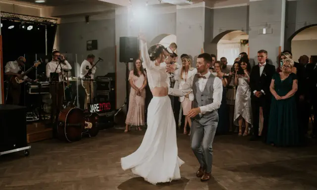 Para młoda tańczy na parkiecie podczas wesela w remizie. W tle zespół gra muzykę, a goście obserwują.