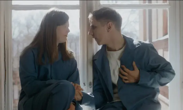 Två unga människor, klädda i blåa arbetskläder, sitter vid ett fönster. Scen från filmen om vipeholmsexperimenten.