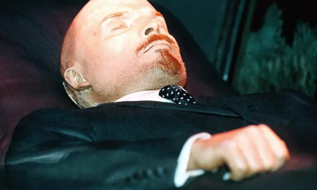 Mumia Lenina w garniturze i z krawatem w kropki, spoczywająca w szklanej gablocie.
