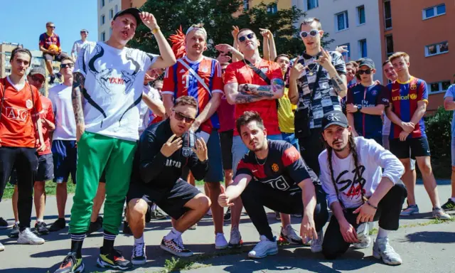 Grupa młodych ludzi w strojach piłkarskich, pozująca do zdjęcia. W tle budynki mieszkalne. To świetny **reto tekst** dla fanów futbolu.