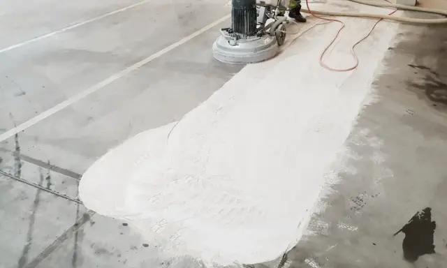Maszyna szlifuje betonową posadzkę, przygotowując ją do naprawy pęknięć. Biały pył pokrywa część podłogi.