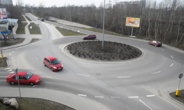 Rowerzysta na rondzie, otoczony przez czerwone samochody i drzewa.