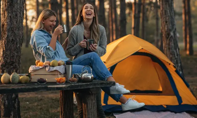 Dwie kobiety śmieją się na kempingu, siedząc przy stole z owocami i napojami. Obok rozstawiony jest żółty namiot.