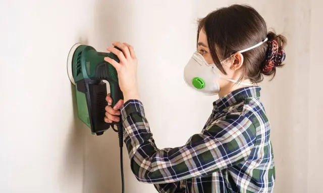 Mujer con mascarilla lijando una pared con una lijadora eléctrica.