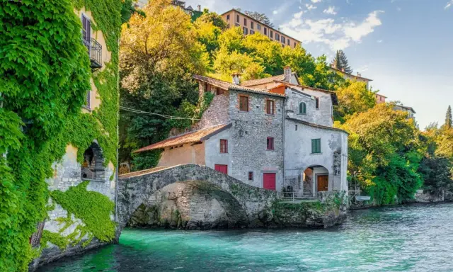 Antica casa in pietra con edera su un lago turchese, un ponte ad arco e colline verdi sullo sfondo, un angolo da fiaba dei castelli Lombardia.