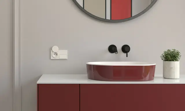 Nowoczesna instalacja elektryczna w łazience: białe gniazdko z klapką i włącznik obok bordowej umywalki.