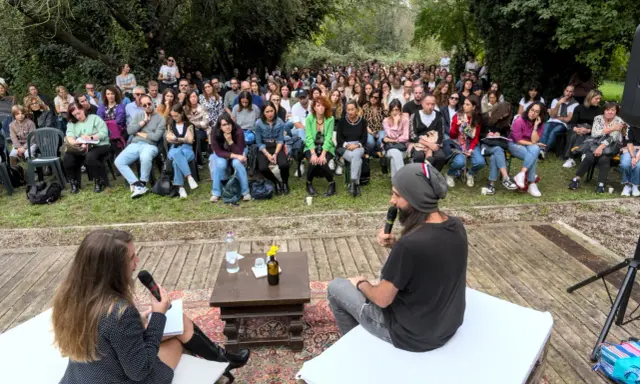 Un pubblico attento ascolta un dibattito all'aperto. Le parole fluiscono, profonde come il mare, leggere come il cielo.