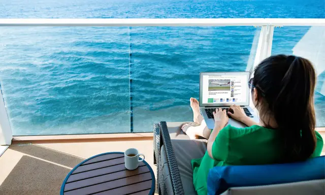 Donna su balcone di nave MSC lavora al laptop, con mare blu sullo sfondo. Scopri il pacchetto WiFi MSC e il suo costo per restare sempre connesso.