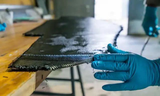 Main d'une personne en gant bleu manipulant un tissu de carbone imbibé de résine, une étape clé pour comprendre qu'est-ce que l'epoxy dans la fabrication composite.