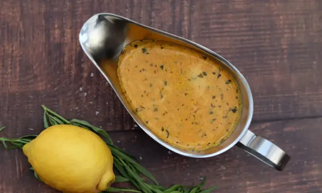 Eine cremige Cafe de Paris Sauce in einer Sauciere, daneben ein Zitrone und Kräuter auf dunklem Holz.