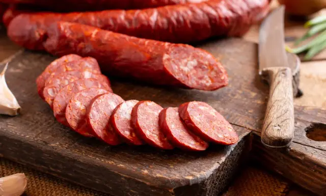 Salami auf einem Holzbrett, geschnitten und im Ganzen. Man kann Salami einfrieren, um sie länger haltbar zu machen. Knoblauch und Messer liegen daneben.