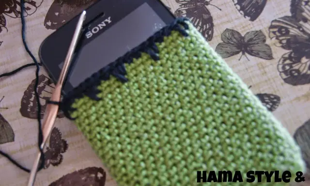 Funda de móvil tejida a crochet, un patrón verde con borde negro. Un gancho y hilo negro están listos para continuar el proyecto.