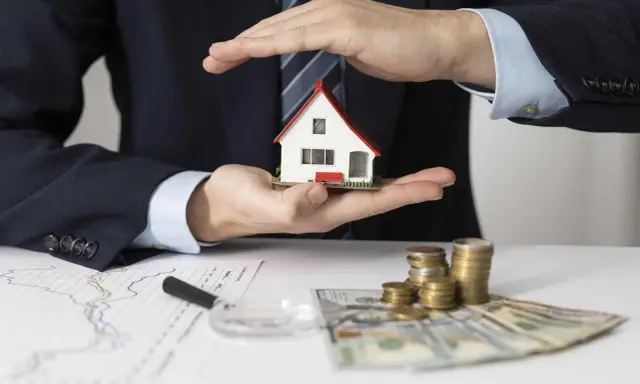 Manos protegen una casa modelo sobre un contrato de alquiler con avalista, junto a dinero y gráficos.