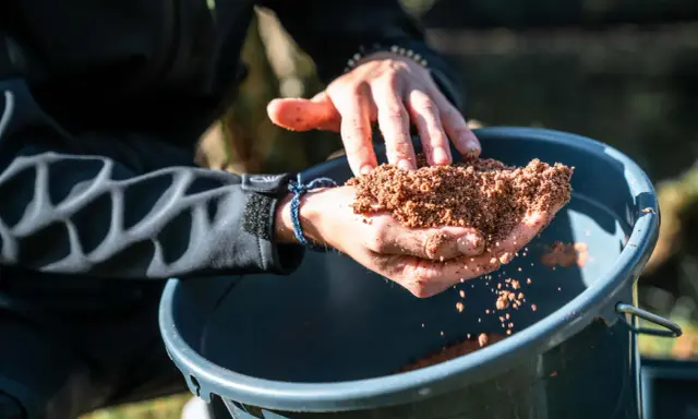 Ręce trzymają zanętę Traper Feeder nad wiaderkiem.