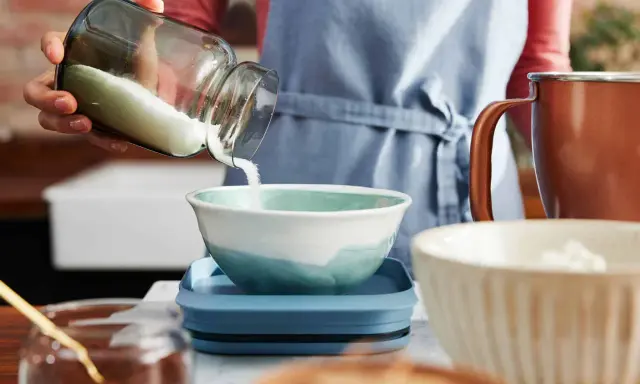 Eine Hand schüttet Mehl aus einem Glas in eine Schüssel. Die Frage, wie viel Gramm Mehl in eine Tasse passen, wird hier visuell thematisiert.