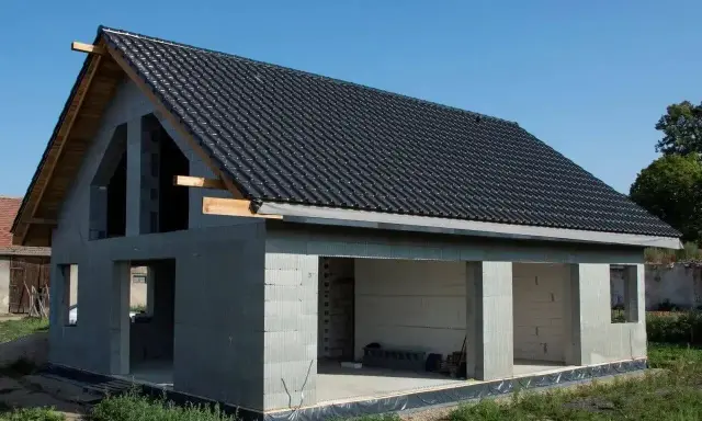 Budowa domu stan surowy: szary beton, czarny dach, drewniane elementy konstrukcyjne. Widać otwory okienne i drzwiowe.