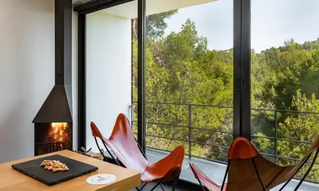 Chimenea encendida y sillones de cuero frente a un balcón con vistas a un bosque frondoso, ideal para el turismo interior España.