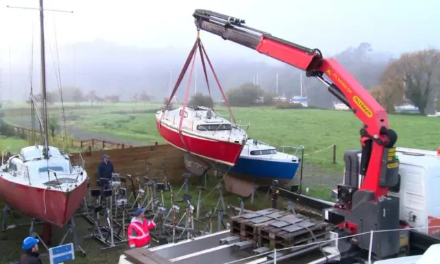 Grue soulevant un bateau rouge pour son recyclage. Un autre bateau bleu est posé sur des supports. L'opération de recyclage bateau se déroule dans un paysage brumeux.