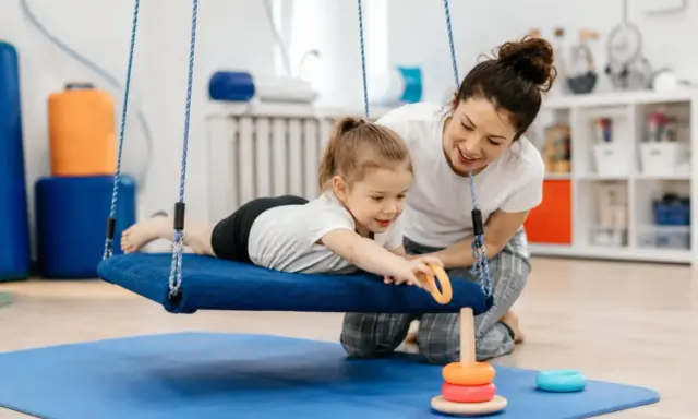 Terapia integracji sensorycznej: dziewczynka na huśtawce z terapeutką, która pomaga jej sięgnąć po zabawkę.