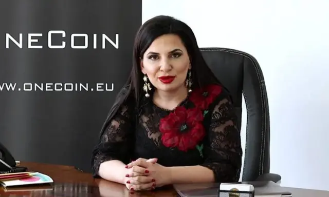 Rupert Ignatov, w czarnej koronkowej sukni z czerwonymi kwiatami, siedzi przy biurku.