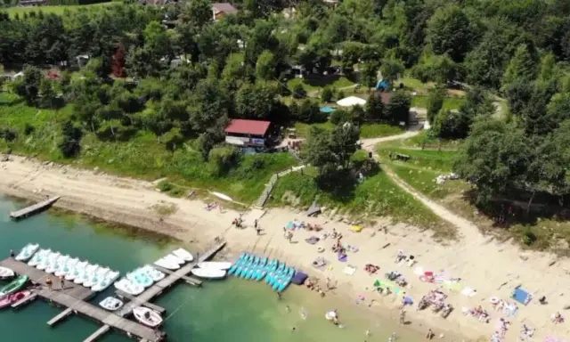 Słoneczna plaża w Polańczyku tętni życiem. Ludzie odpoczywają na piasku, a w wodzie pływają rowery wodne i kajaki.