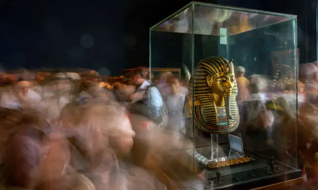 Goldene Maske des Pharao Tutanchamun in einer Vitrine, umgeben von verschwommenen Besuchern.