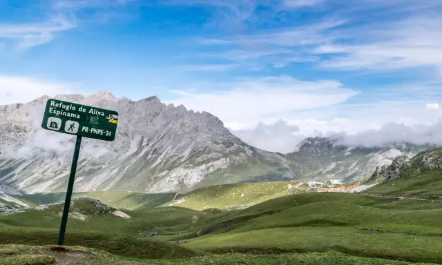 Wanderwegschild weist den Weg zum Refugio de Aliva und Espinama in den **Lagos de Covadonga**. Grüne Hügel und zerklüftete Berge unter blauem Himmel.