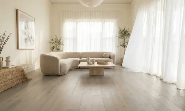 Un salon lumineux avec un canapé incurvé beige, une table basse en pierre et un sol en bois gris clair. Le beige et le blanc s'accordent parfaitement avec le gris clair.