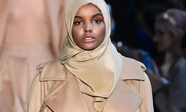 Halima Aden walks the runway in a stylish beige coat and hijab, exuding confidence and grace.
