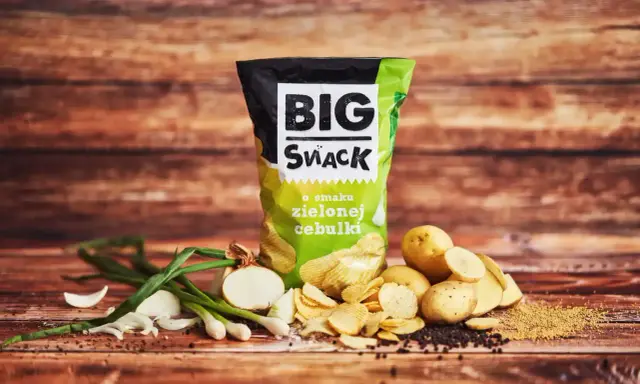 Reklamowa fotografia paczki chipsów "Big Snack" o smaku zielonej cebulki, z ziemniakami i cebulką w tle.