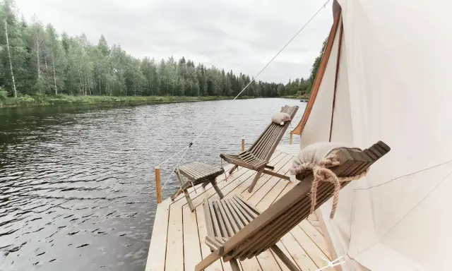 Två träfåtöljer och ett litet bord på en brygga vid en sjö, med skog i bakgrunden. En del av ett glampingtält syns. Upplev glamping Norrbotten.