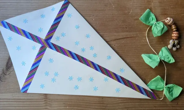 Ein selbstgebastelter Drachen mit Schneeflocken und bunten Bändern, perfekt für basteln mit Kindern ab 7 Jahre. Daneben eine Schnur mit grünen Schleifen und Holzperlen.