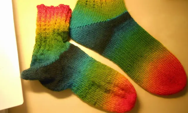 Två regnbågsfärgade, handstickade sockor i ett stycke. En större och en mindre, med färger som går från rosa och gult till grönt och blått.