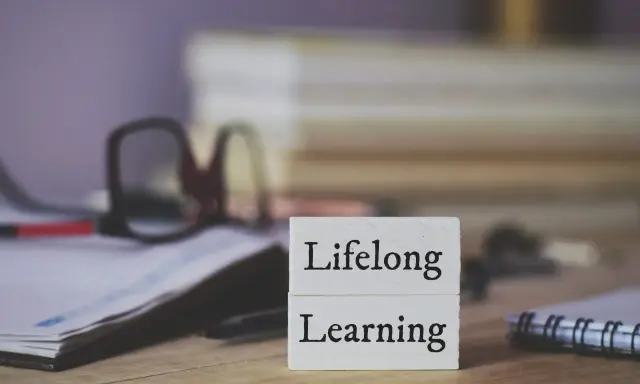Dwa białe klocki z napisami "Lifelong" i "Learning" symbolizują uczenie się przez całe życie. W tle okulary i notatnik.