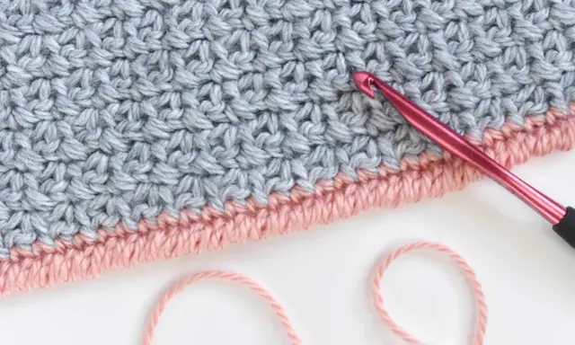 Detalle de tejido a crochet con punto cangrejo en rosa y gris. Una aguja rosa está lista para continuar el trabajo.