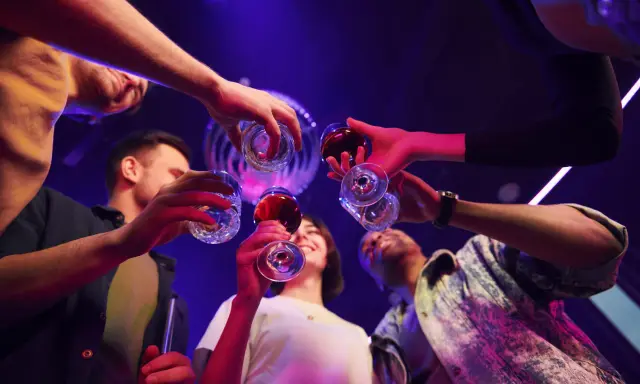 Grupa przyjaciół wznosi toast, świętując weekend. W tle dyskotekowe światła, sugerujące zabawę i być może początek weekendowego alkoholizmu.