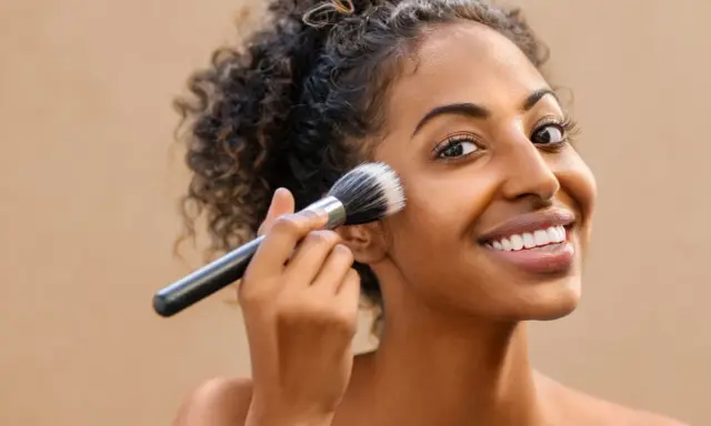 Donna sorridente applica il fondotinta con un pennello. La sua pelle luminosa è il risultato di cosmetici nichel free.