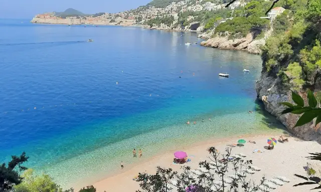 Malownicza zatoka z turkusową wodą i kamienistą plażą. W oddali widać mury Starego Miasta Dubrownik. Idealne plaże Dubrownik na relaks.