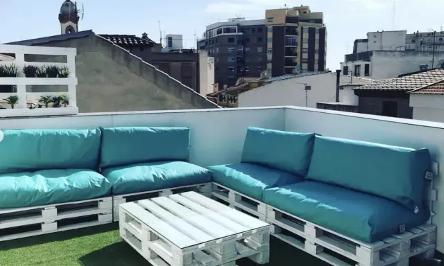 Sofá y mesa de centro hechos con palets blancos, con cojines turquesas, en una terraza urbana. ¡Una idea genial de como hacer un sofa con palets!